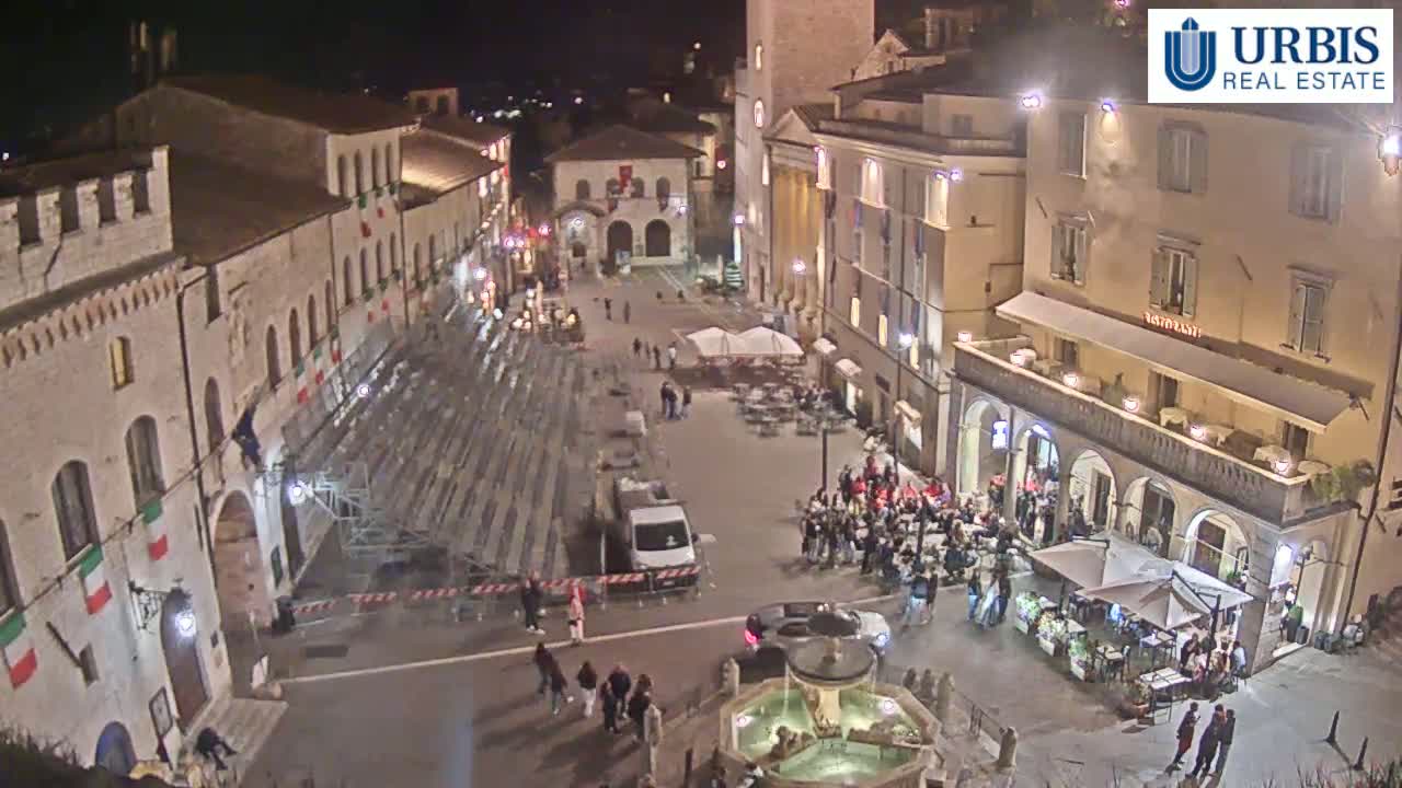 A nighttime aerial view of a town square with several buildings, a fountain, temporary seating, and a small crowd of people.