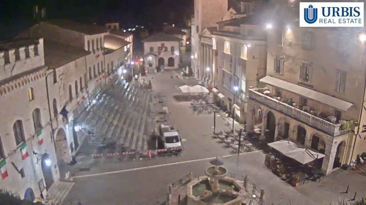 A nighttime, high-angle view of a town square with empty seating, a fountain, and buildings illuminated under artificial light.