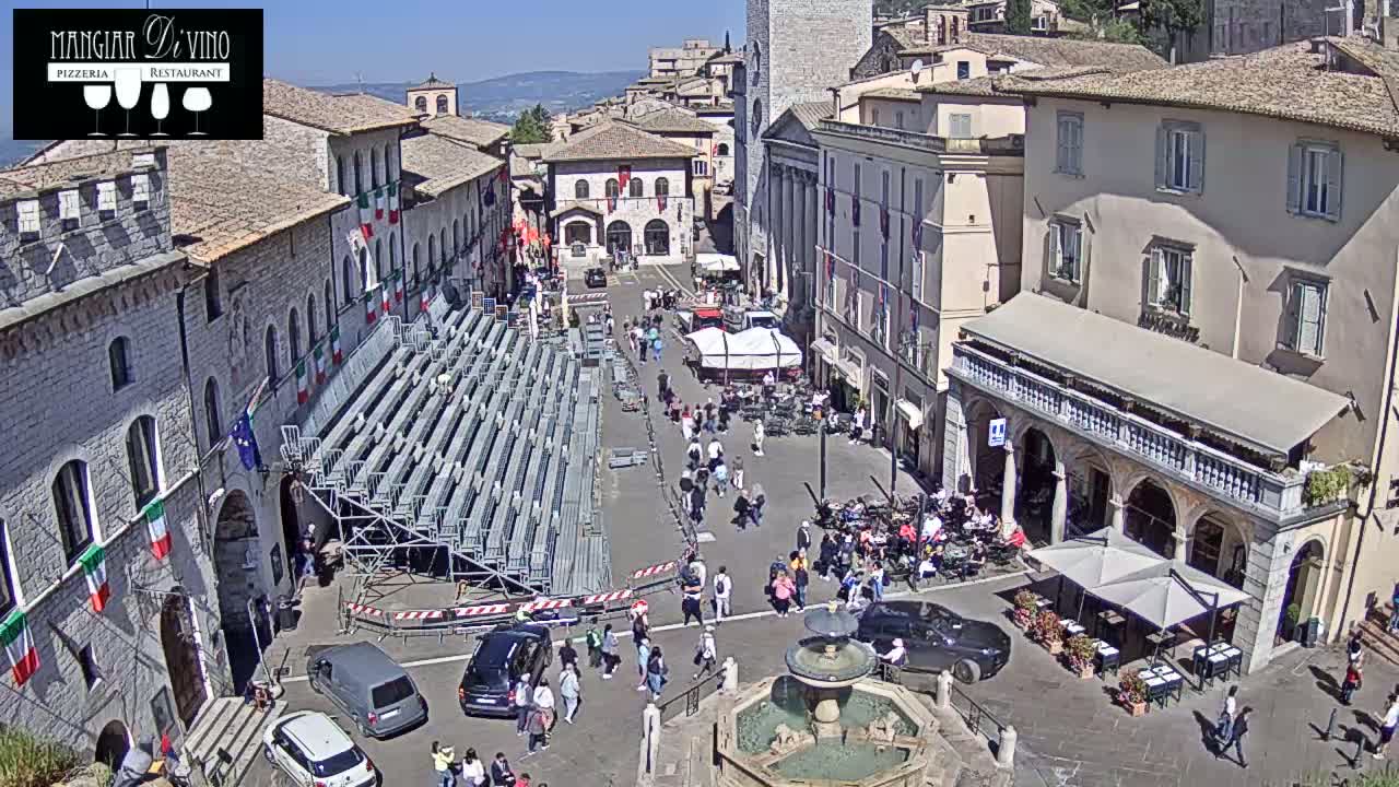 A sunny outdoor town square scene shows a large temporary seating structure, several people walking around, cars parked, and buildings with Italian flags hanging from them.