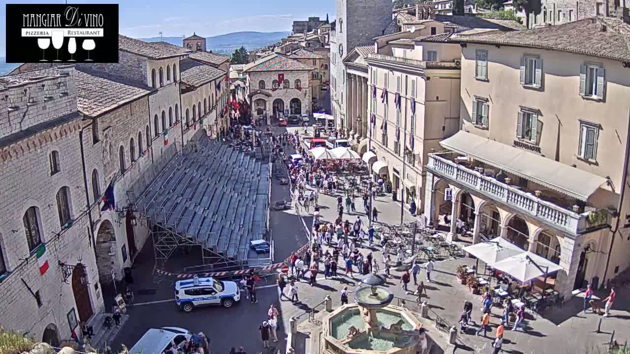 A sunny day in a town square shows a crowd of people gathered around a fountain, near temporary bleachers and outdoor cafes, with buildings lining the square.