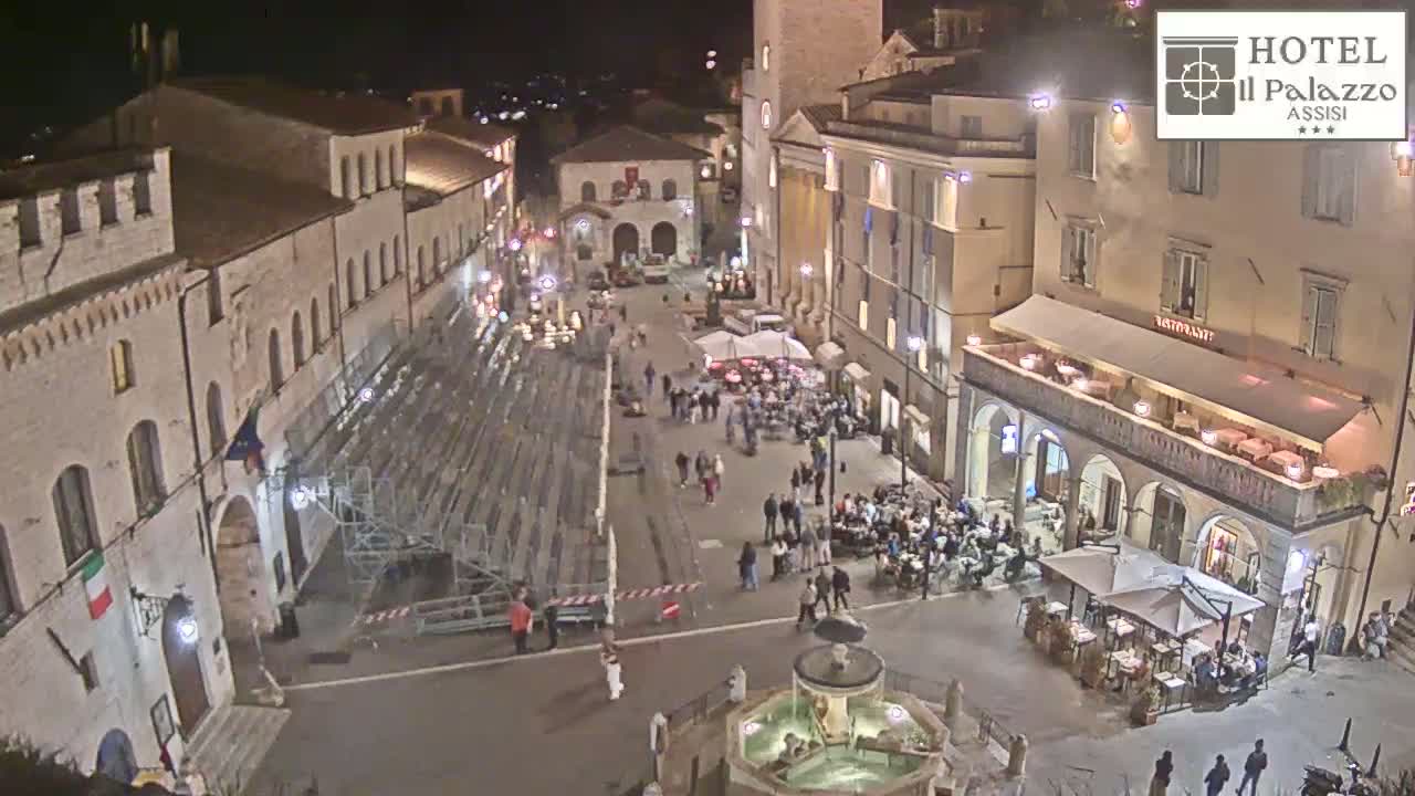 A nighttime aerial view of a town square, with people gathered around outdoor seating at restaurants and a fountain in the center, under clear skies.