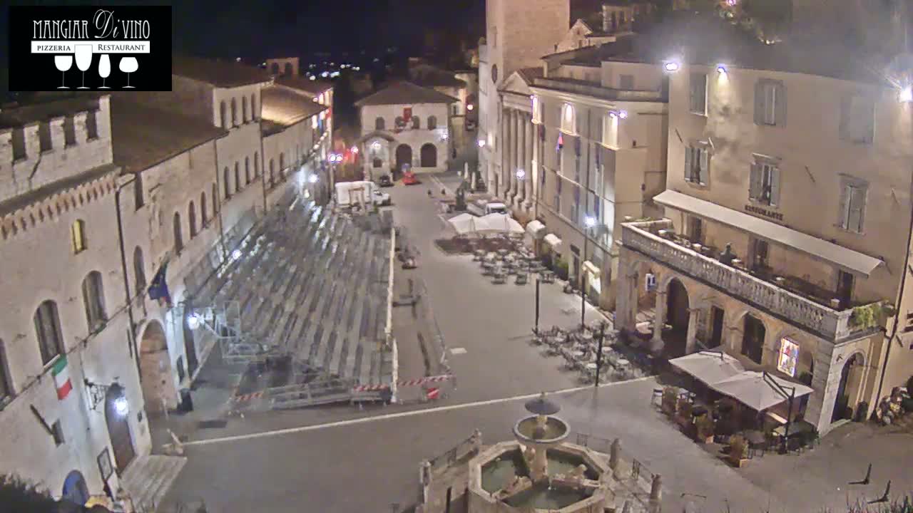 A nighttime, high-angle view of a town square with temporary seating, several buildings, and a fountain.