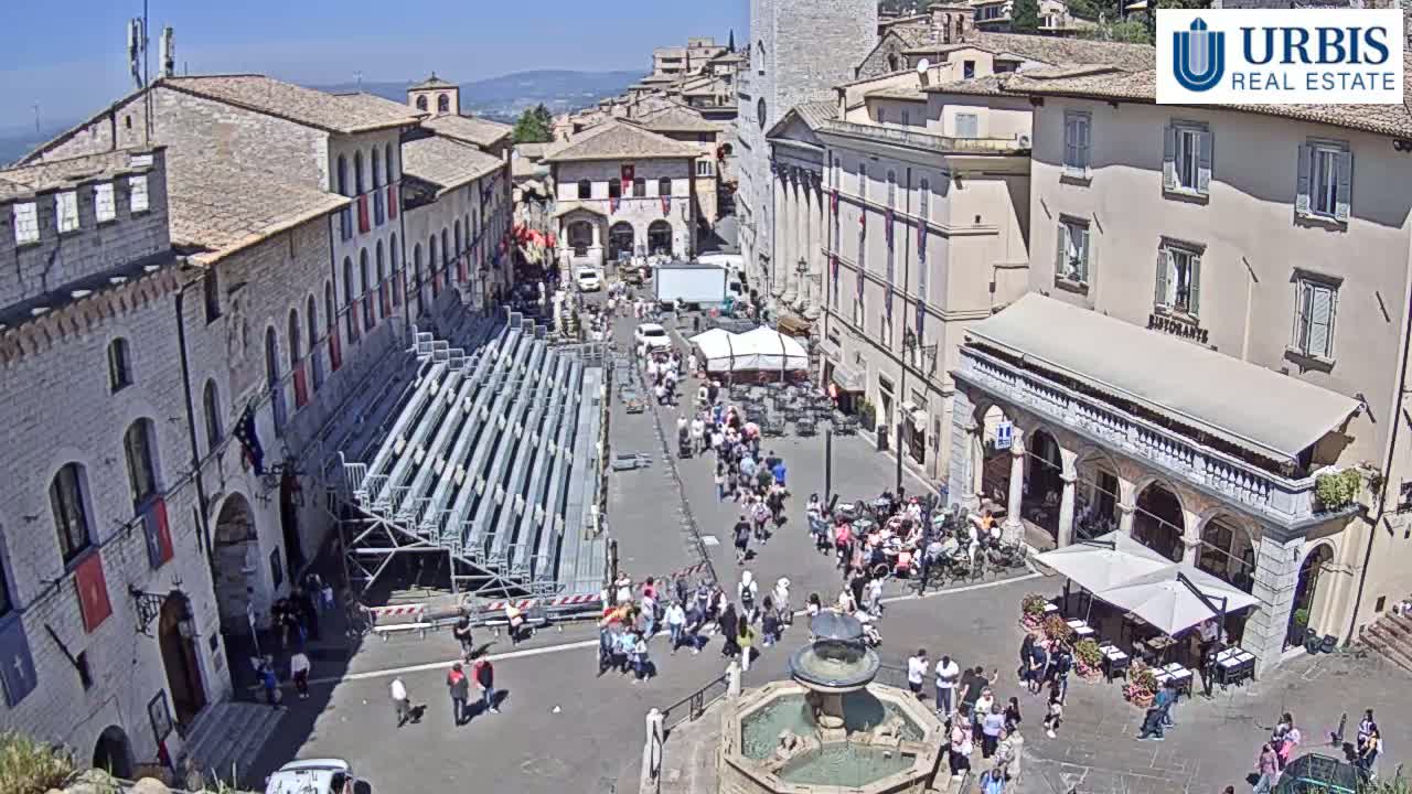 A sunny town square, filled with people and temporary seating, is surrounded by historic buildings under a clear blue sky.