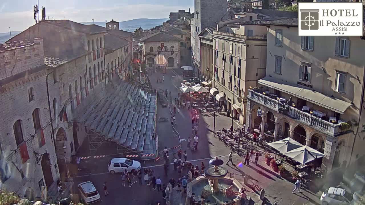 A sunny town square, filled with people and outdoor seating, features a large set of bleachers and a fountain, surrounded by historic stone buildings.