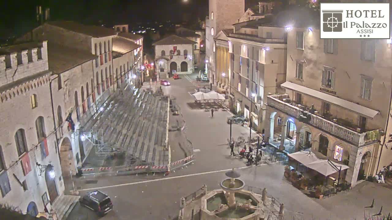 A nighttime, high-angle view of a town square with stone buildings, temporary seating, a fountain, and a few people.