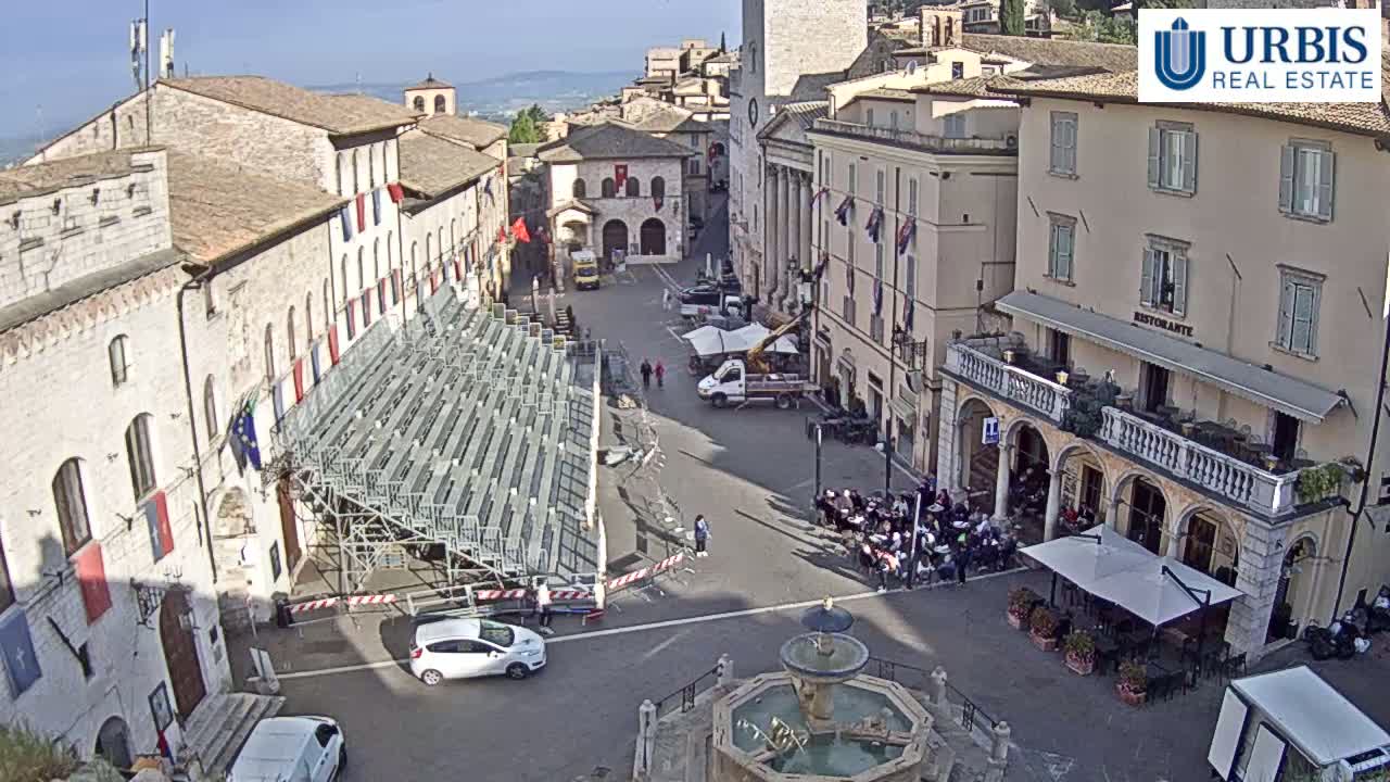 A town square with stone buildings, a temporary seating structure, a fountain, and outdoor seating at a restaurant under sunny skies.
