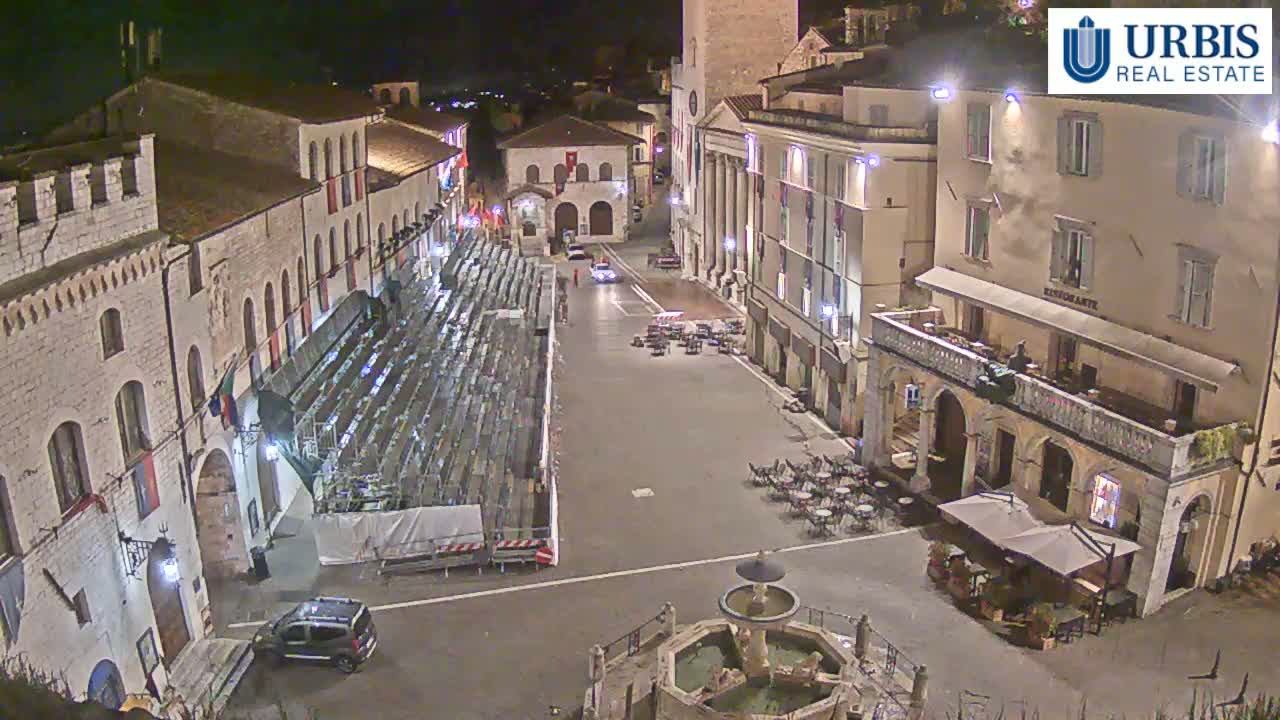 An empty town square at night, illuminated by streetlights, features a fountain, outdoor seating areas, and a large set of empty bleachers set against the backdrop of several multi-story buildings.