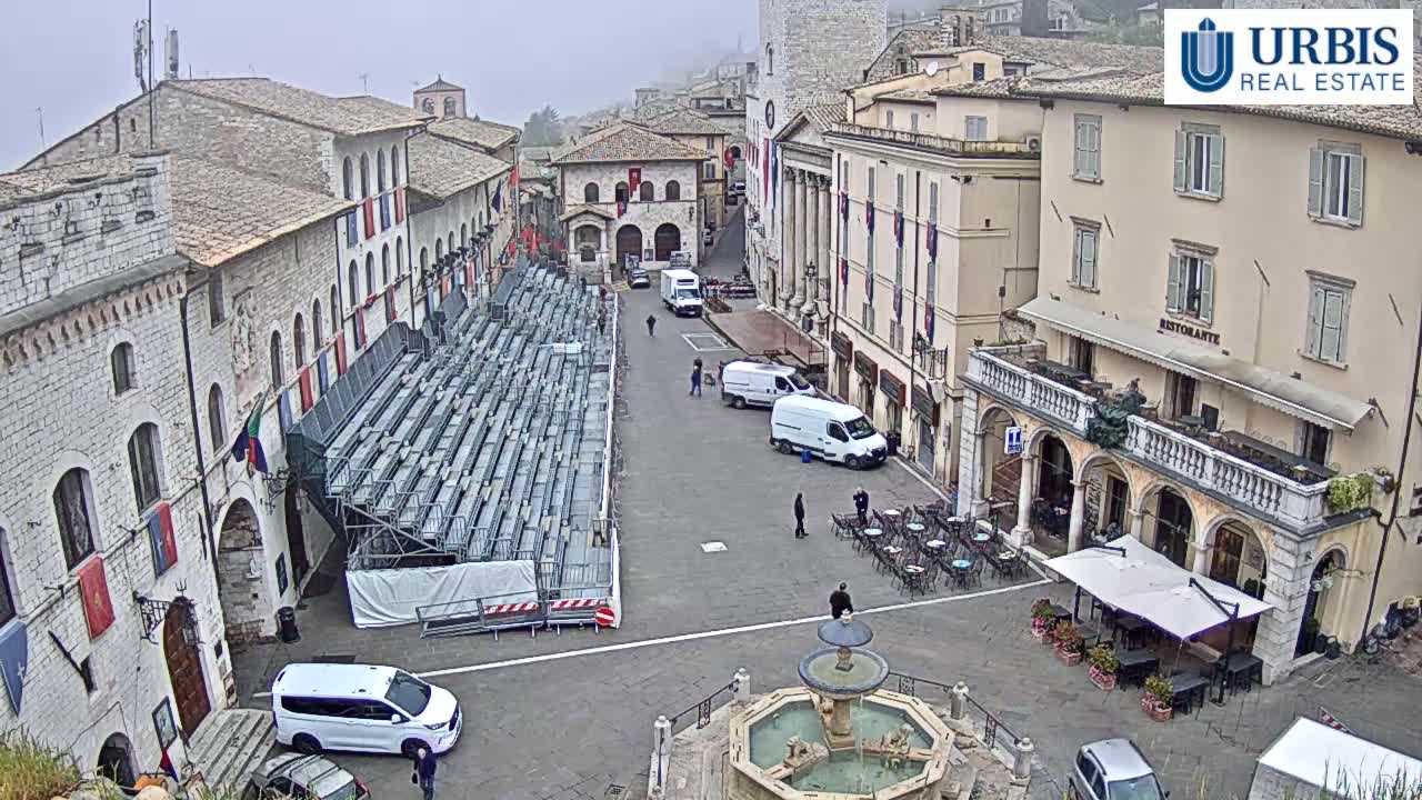 A mostly empty town square, under an overcast sky, features temporary bleachers, several parked vans, a fountain, and outdoor seating at a restaurant.