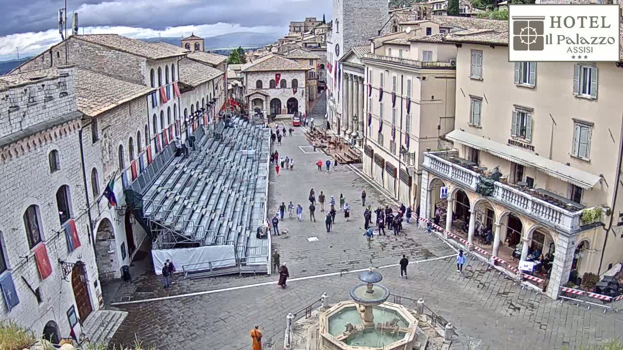 A town square with temporary bleachers, a fountain, and several buildings under an overcast sky.