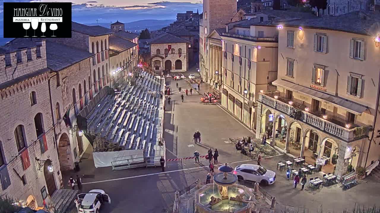 A twilight view of an empty town square with temporary bleachers, a fountain, and several people sparsely scattered throughout.