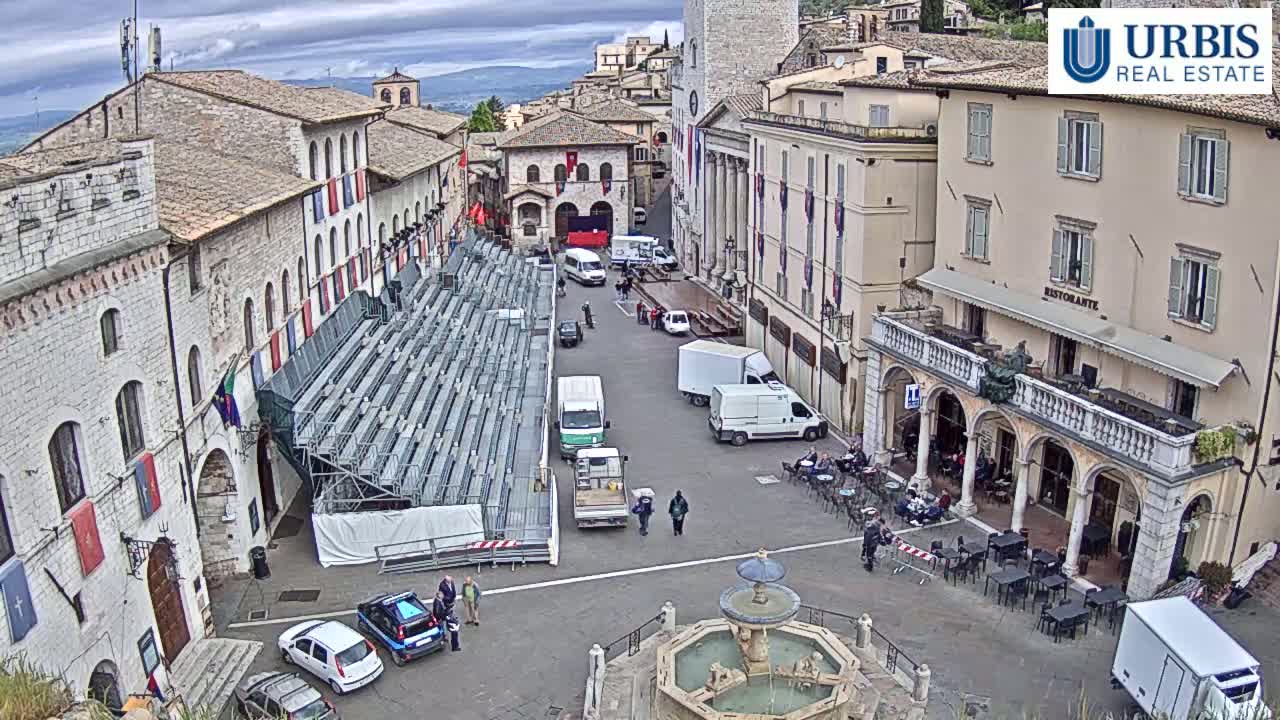A town square with temporary bleachers, vehicles, and a fountain, under an overcast sky.