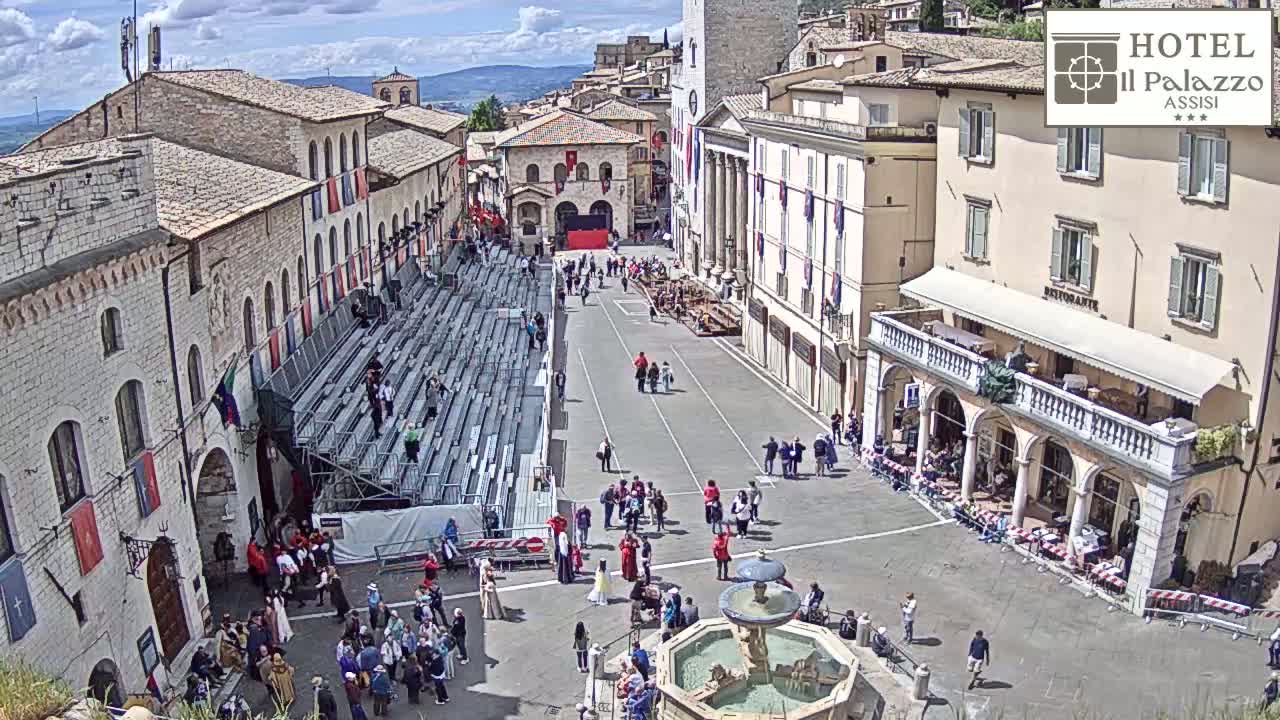 A sunny town square, featuring a large temporary seating structure, a fountain, and several historic-looking buildings.