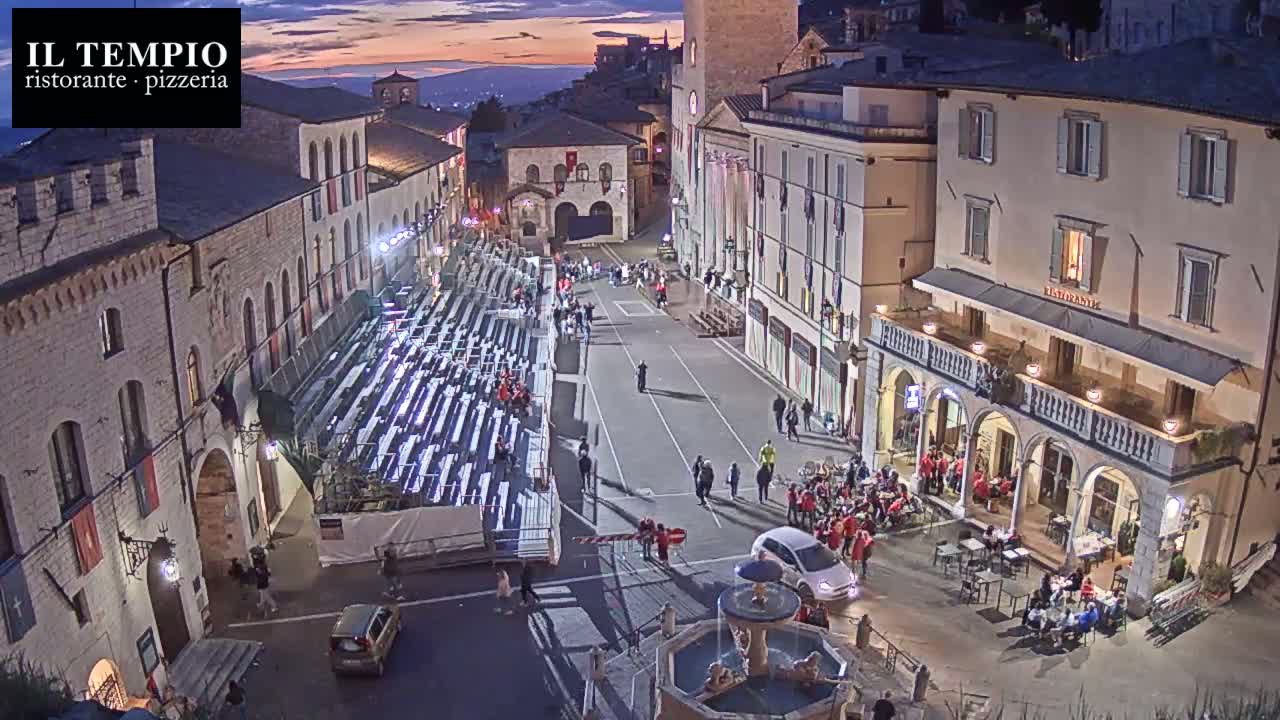 A town square at dusk is filled with people, temporary seating, and a fountain, with buildings lining the sides.