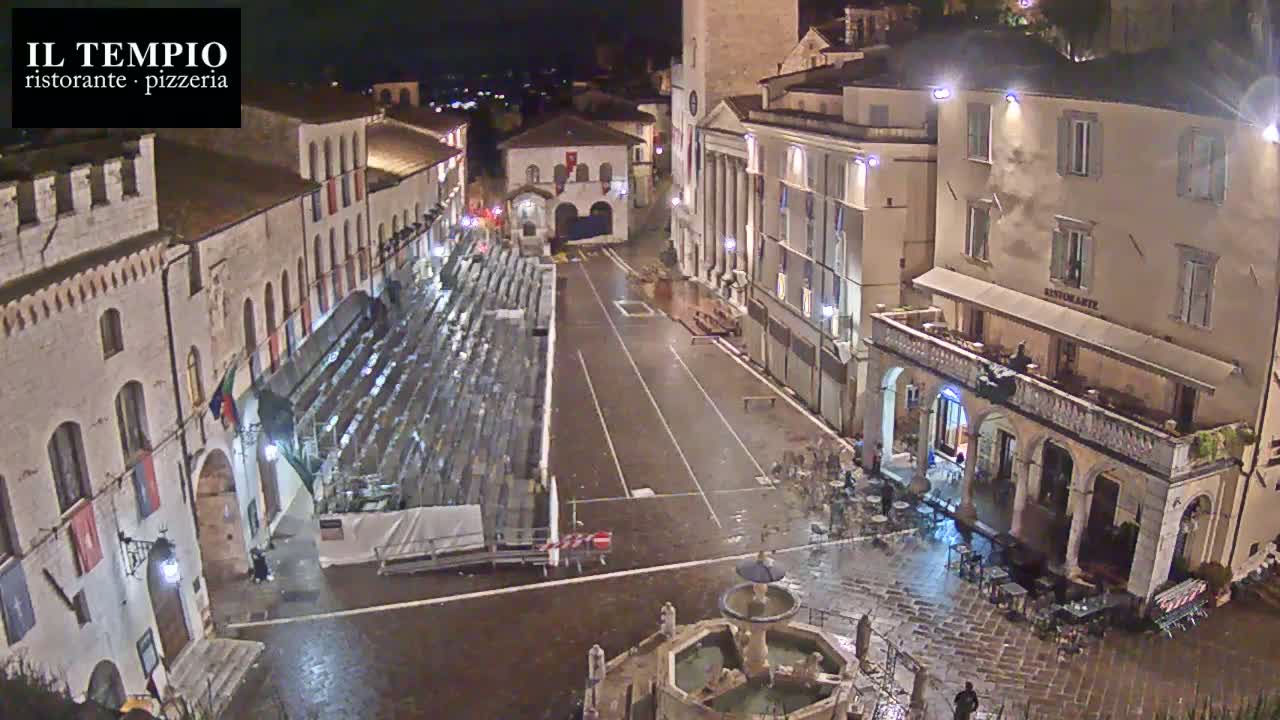 A wet, nighttime view of a town square with empty bleachers, a fountain, and several buildings, one of which is a restaurant.