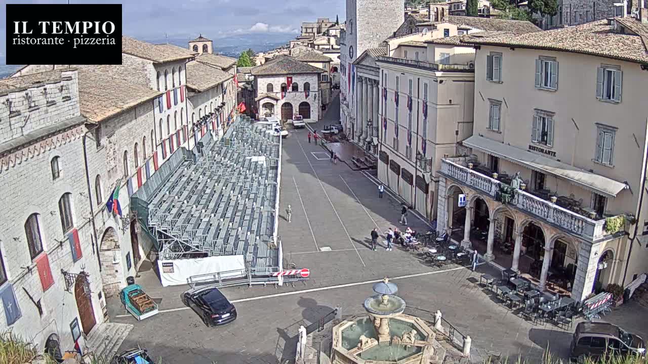 A town square with a fountain, temporary seating, and several buildings, under sunny conditions.