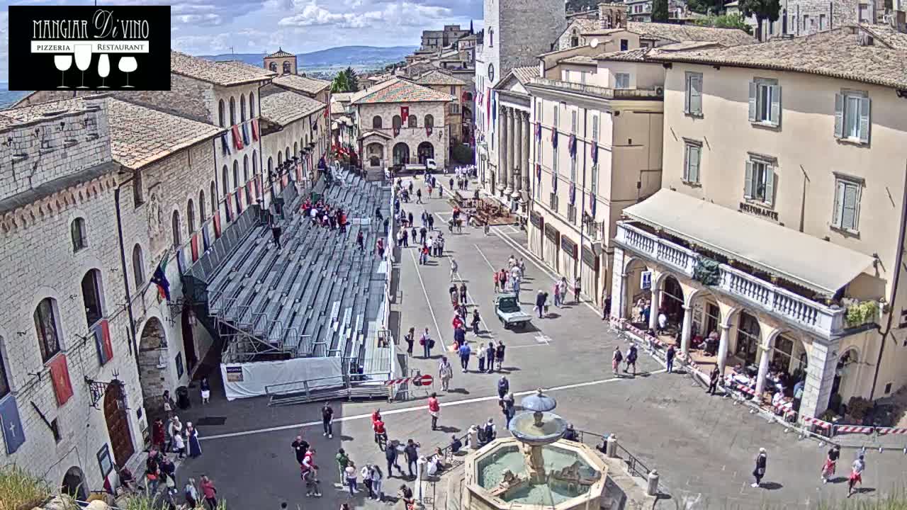 A town square with temporary bleachers, a fountain, and many people milling about on a partly cloudy day.