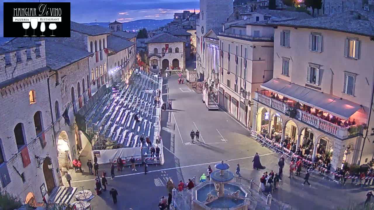 An outdoor town square at dusk, featuring empty bleachers, a fountain, and several people walking around buildings.