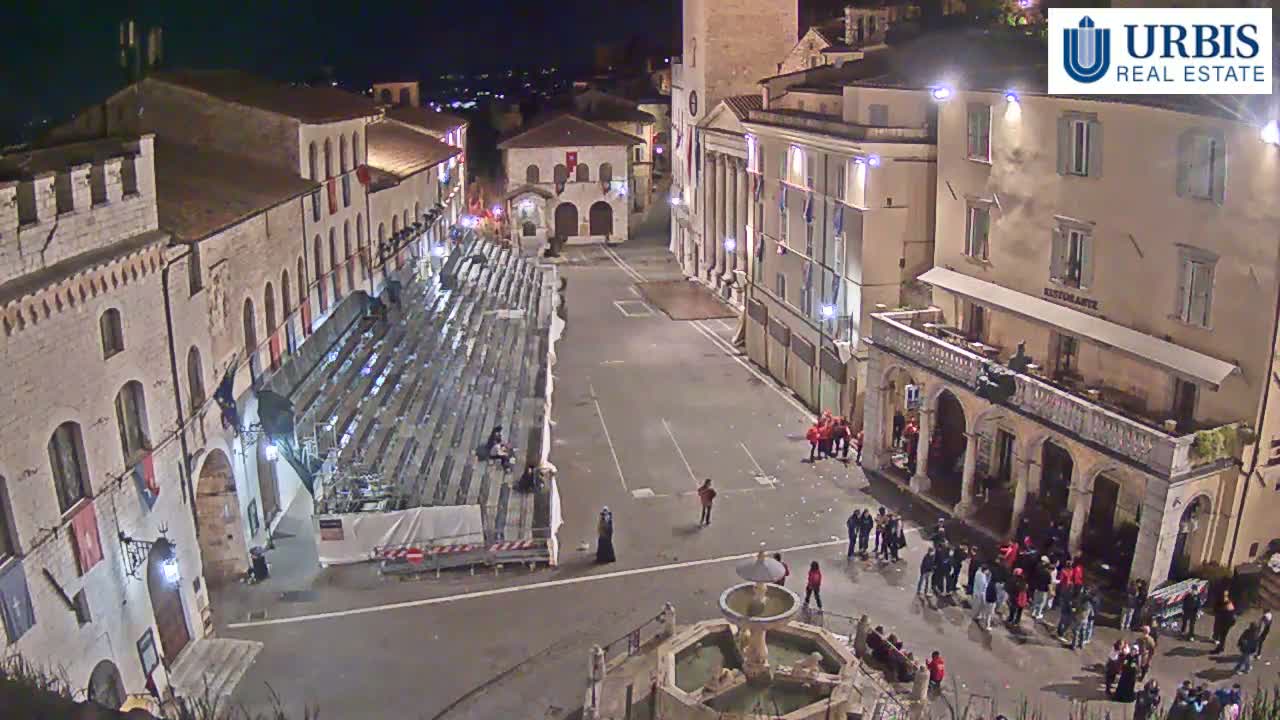 A nighttime aerial view shows a town square with empty bleachers, a fountain, and several people scattered throughout the area.
