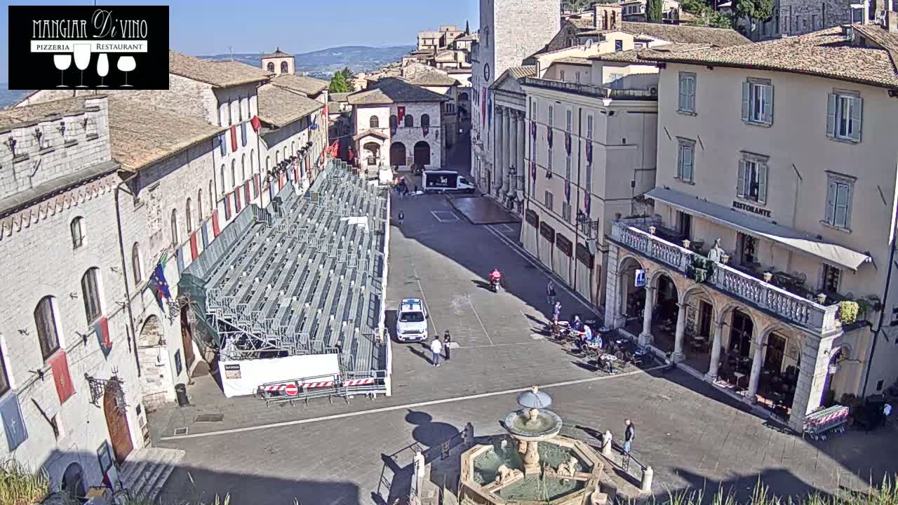 A mostly empty town square with a temporary stage, fountain, and several buildings under a sunny sky.