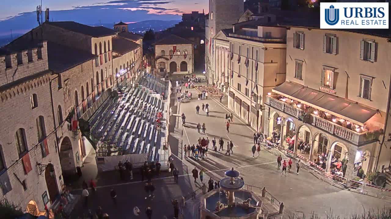 An elevated view of a town square at twilight shows stone buildings, temporary seating, and many people gathered in the square around a fountain.