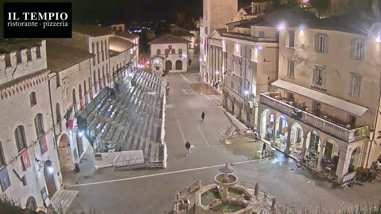A nighttime, high-angle view shows a town square with empty bleachers, a fountain, and several buildings illuminated under the dark sky.