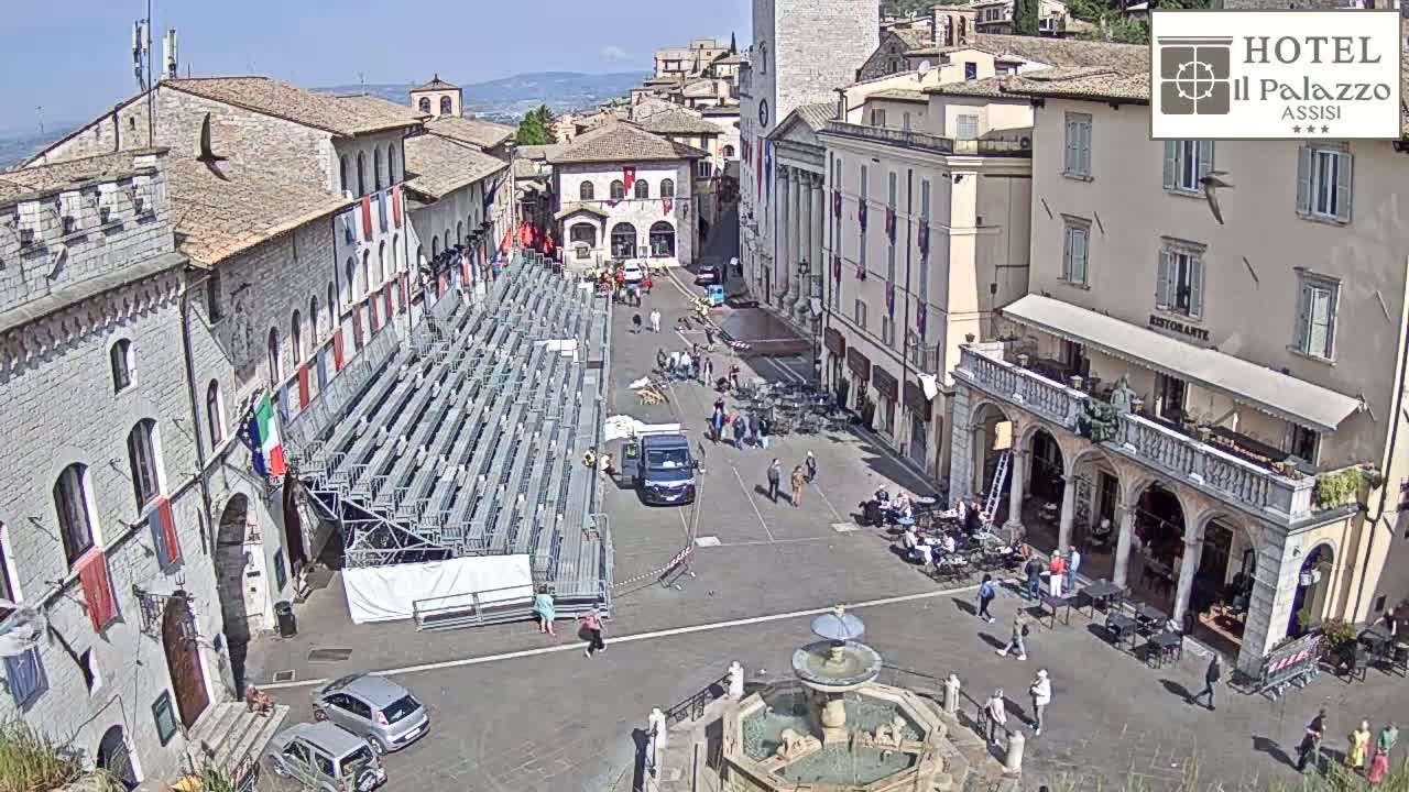 A sunny town square in Italy features a temporary metal bleacher setup, a central fountain, and several stone buildings.