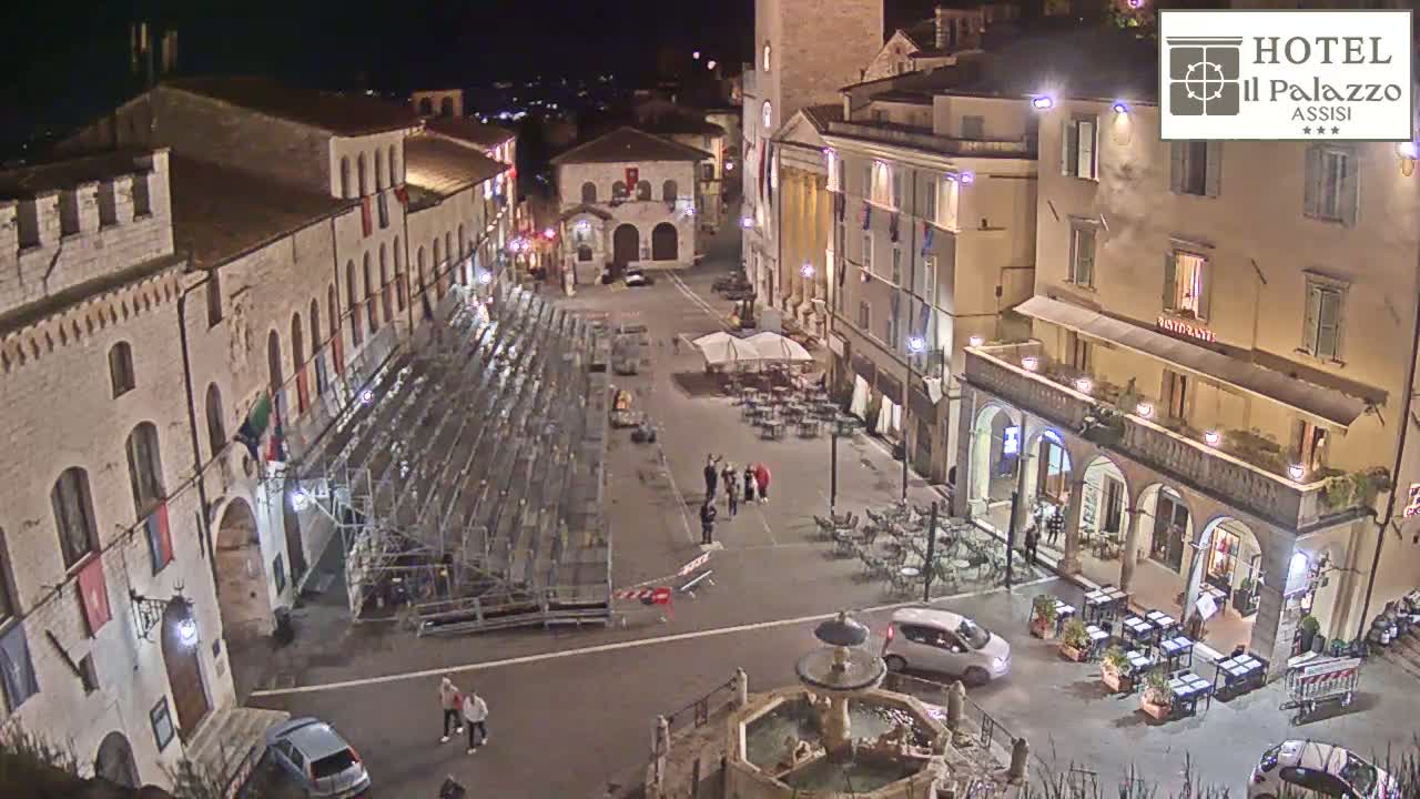 A nighttime aerial view shows an empty town square with a fountain, several buildings, temporary seating, and a few people.