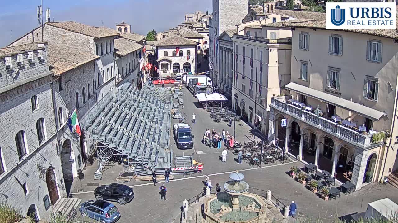 A town square with temporary bleachers, several vehicles, a fountain, and outdoor seating at restaurants, under sunny skies.