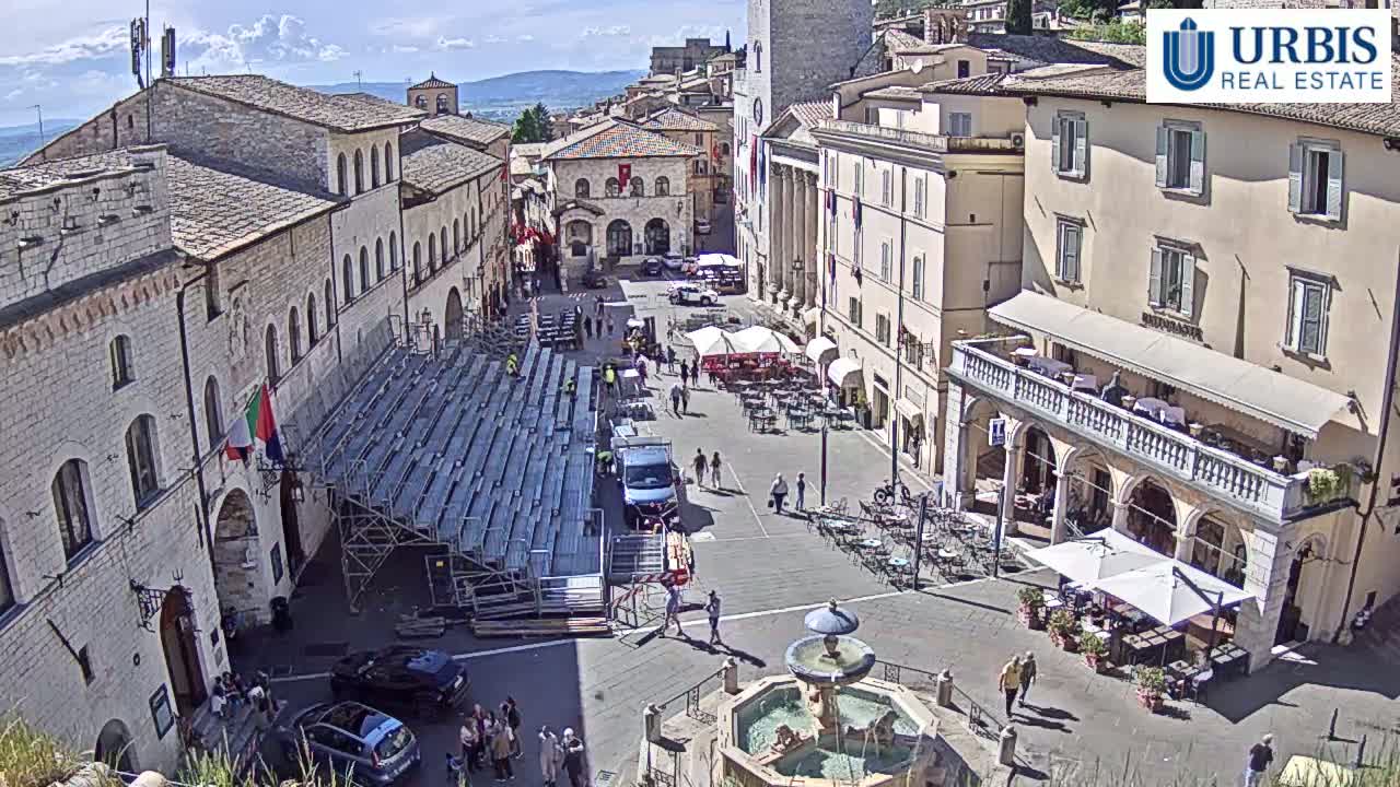A sunny town square in a European-style town, with temporary bleachers set up, outdoor seating areas at cafes, a fountain, and several people walking around.