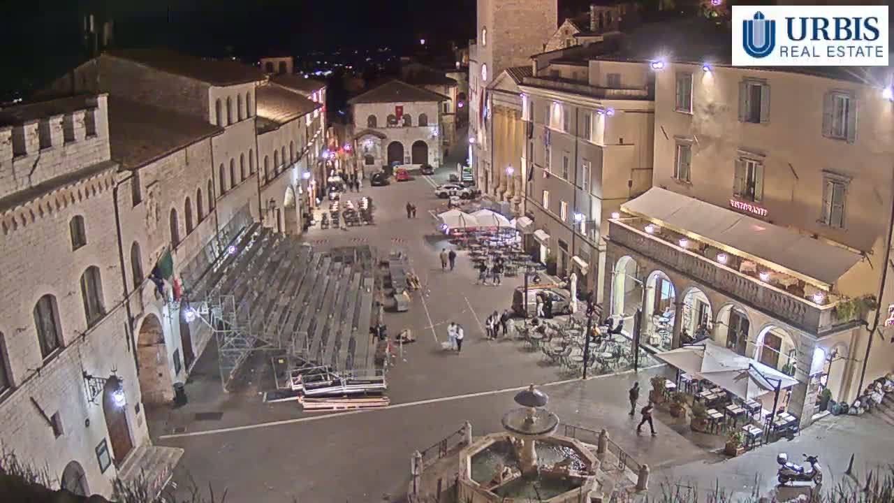 A nighttime view of a town square with several buildings, outdoor seating areas, a fountain, and empty bleachers.
