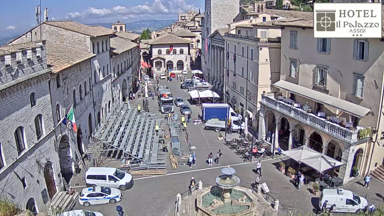 A sunny day reveals a town square in Italy, filled with people, vehicles, temporary seating, and a fountain, surrounded by historical stone buildings.
