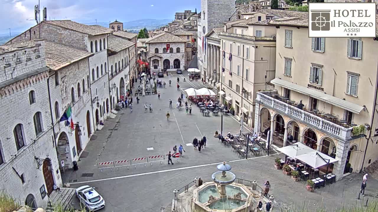 A mostly sunny town square in Italy, filled with people, outdoor seating at restaurants, a central fountain, and buildings in varying shades of tan and light brown.