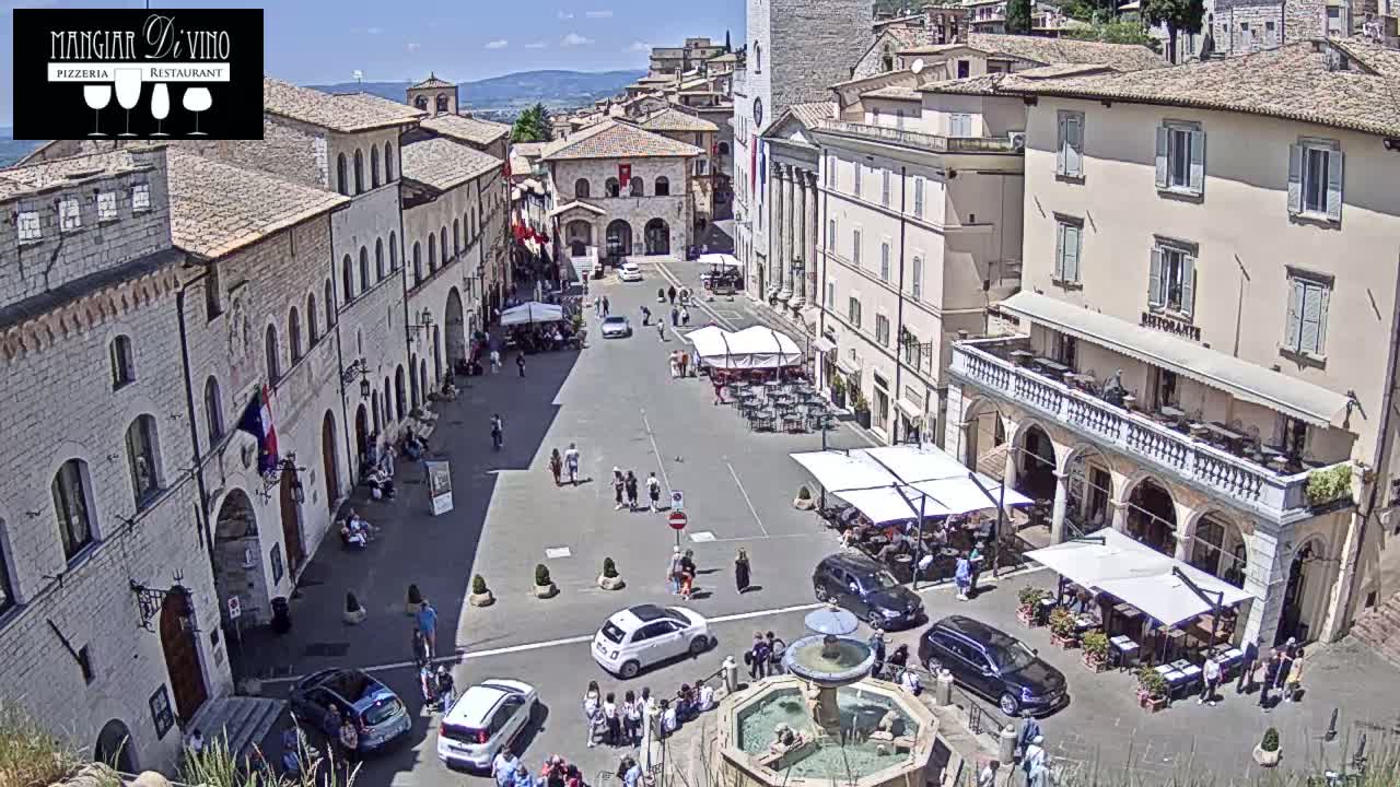 A sunny day in a town square with several buildings, outdoor seating at restaurants, cars parked, people walking around, and a fountain in the center.