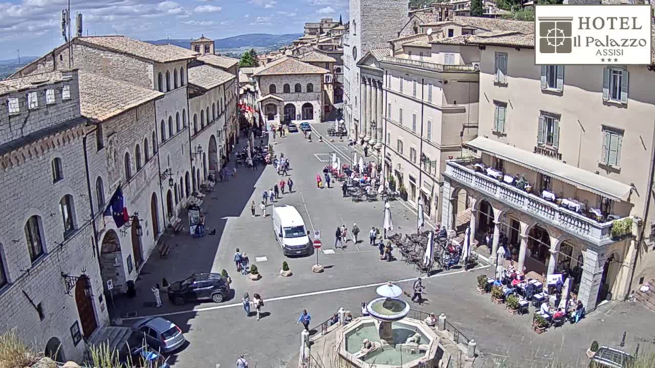 A sunny day in a European town square, showing stone buildings, people walking and sitting at outdoor cafes surrounding a central fountain.