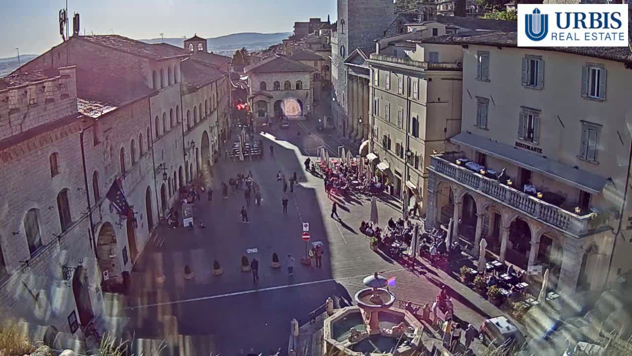 A sunlit town square with several stone buildings, outdoor seating areas, and a fountain is seen from above.