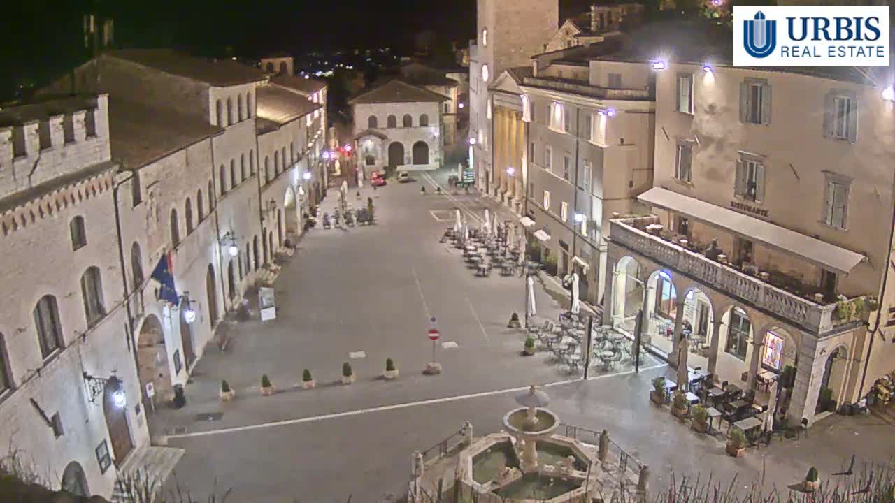 A nighttime, high-angle view of a town square with several buildings, a fountain, and outdoor seating areas at restaurants.