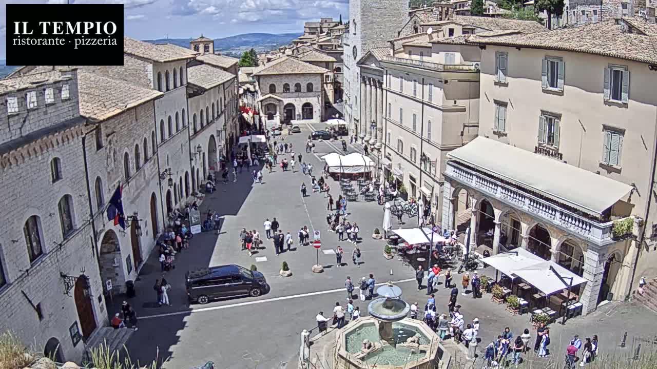 A sunny day in a large, European-style town square with many people, buildings, and a fountain.