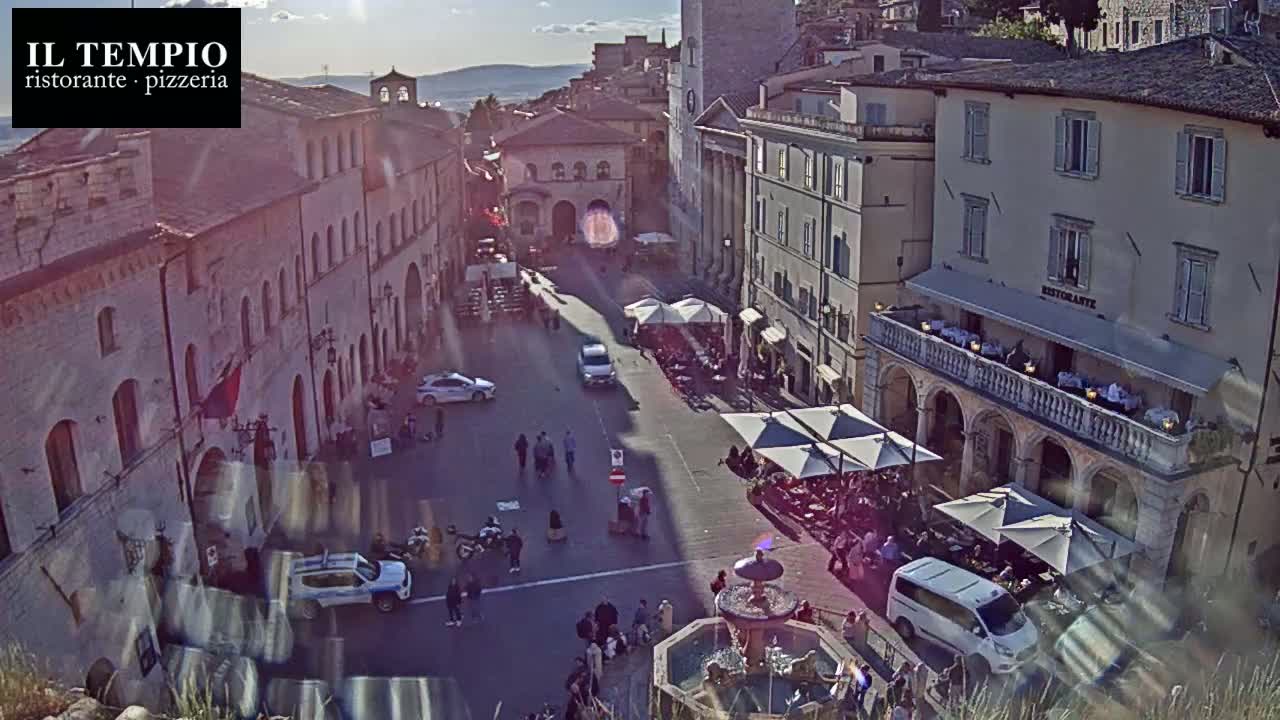 A sunny, high-angle view shows a town square in a European city, filled with people, cars, and outdoor seating at restaurants.