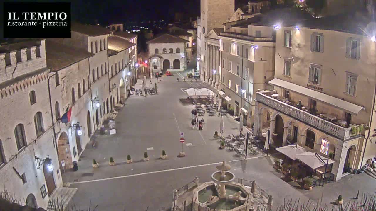A nighttime view of a town square, featuring several buildings, outdoor seating areas, a fountain, and a few pedestrians.