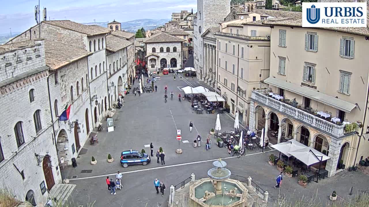 A large, paved town square in a historic Italian city on an overcast day, surrounded by old stone buildings, with a few people and several outdoor cafes.