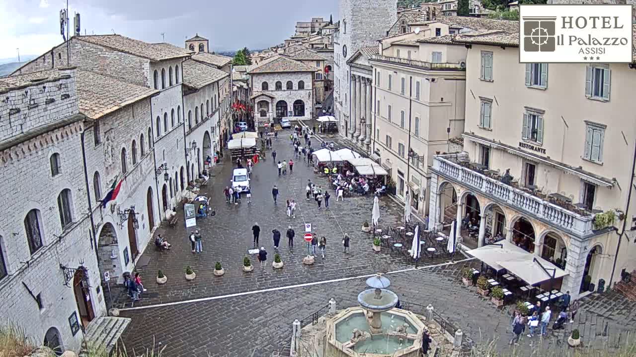 A wet, stone plaza in a European town, surrounded by historic buildings, with people walking around and several outdoor cafes.