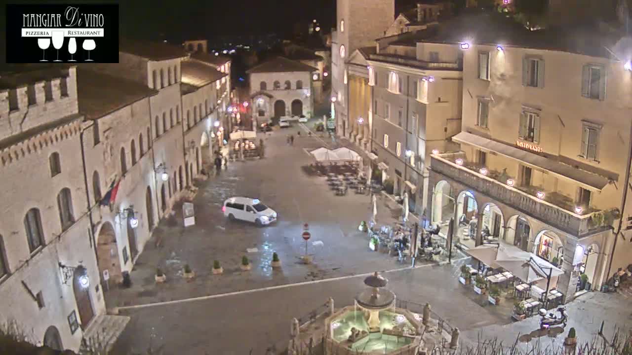 A wet, nighttime view of a town square surrounded by buildings, with a fountain in the center and outdoor seating at various restaurants.