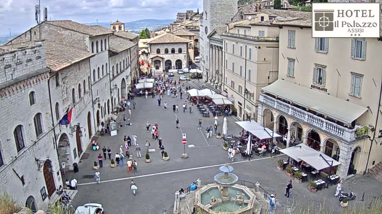A town square in Italy, under partly cloudy skies, features several buildings, a fountain, and numerous people walking and sitting at outdoor cafes.