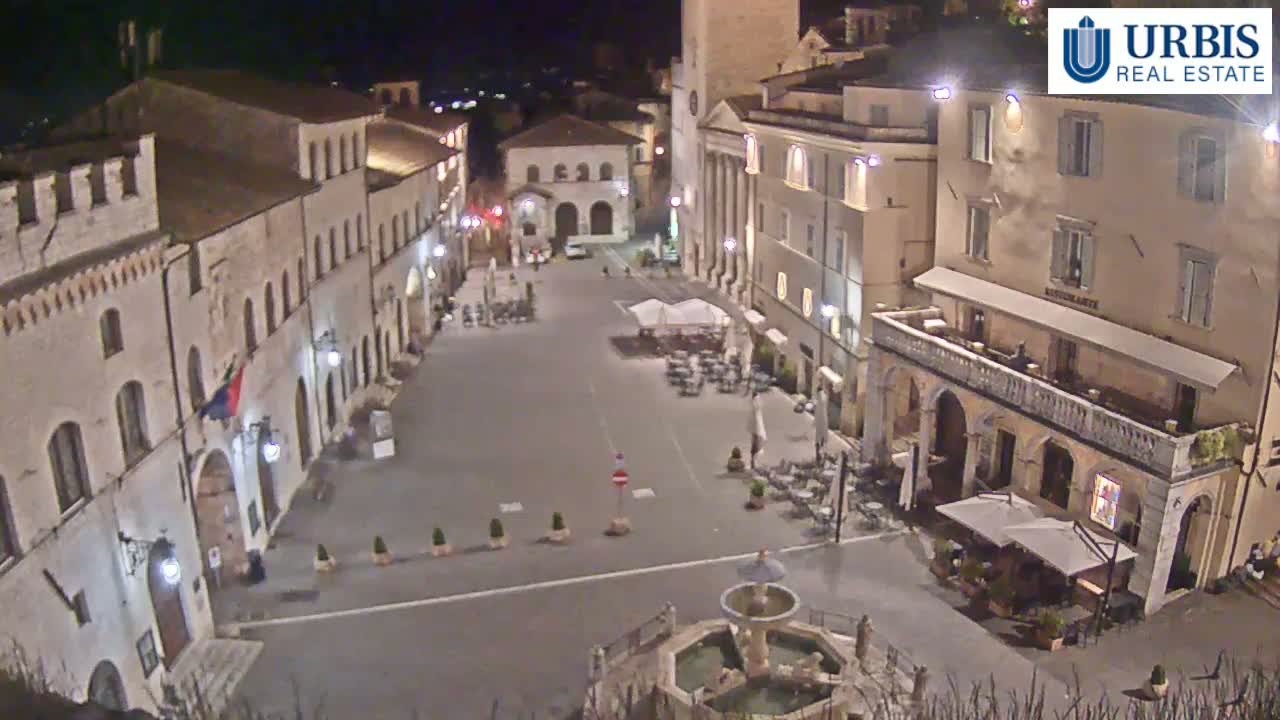 A nighttime, aerial view of a town square in a European-style town, featuring several buildings with outdoor seating areas around a central fountain.