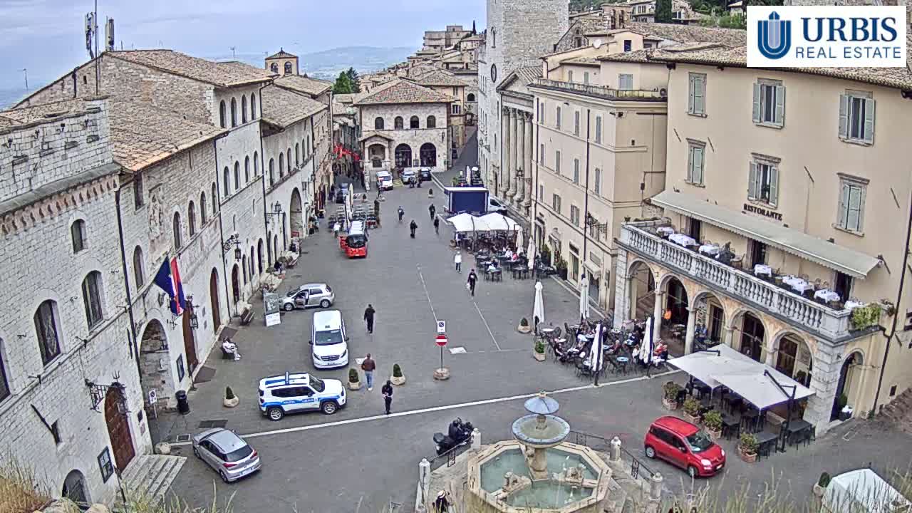 An overcast day reveals a large town square surrounded by stone buildings, with a fountain at its center and several people and cars scattered throughout.