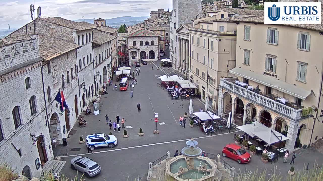 An overcast day reveals a town square in a European city, filled with people at outdoor cafes surrounding a fountain, with cars parked along the edges and buildings lining the square.