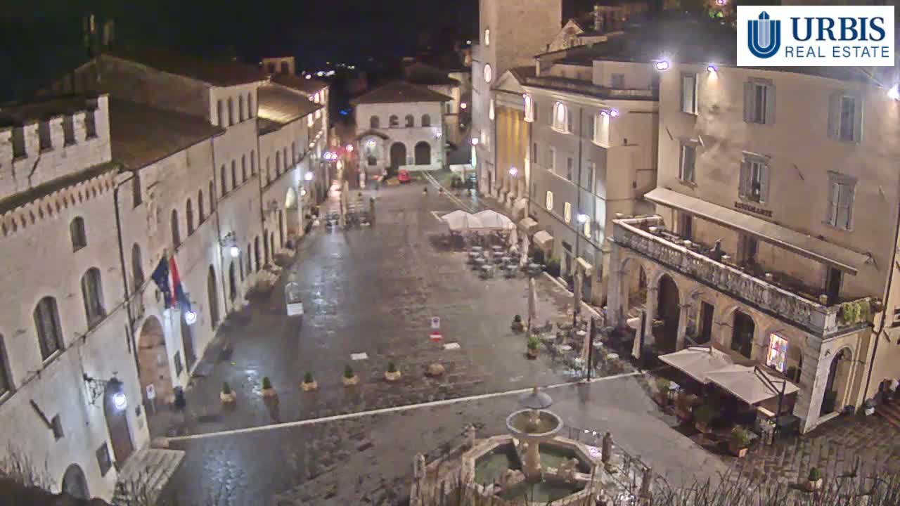 A wet, empty town square at night, surrounded by old stone buildings with outdoor seating areas at some of the buildings.