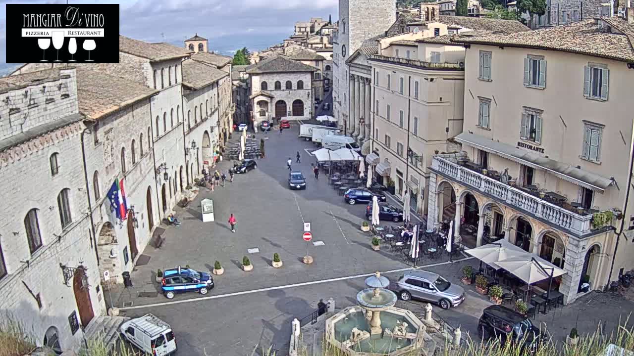An aerial view of a town square with several buildings, cars, a fountain, and outdoor seating areas under partly cloudy skies.