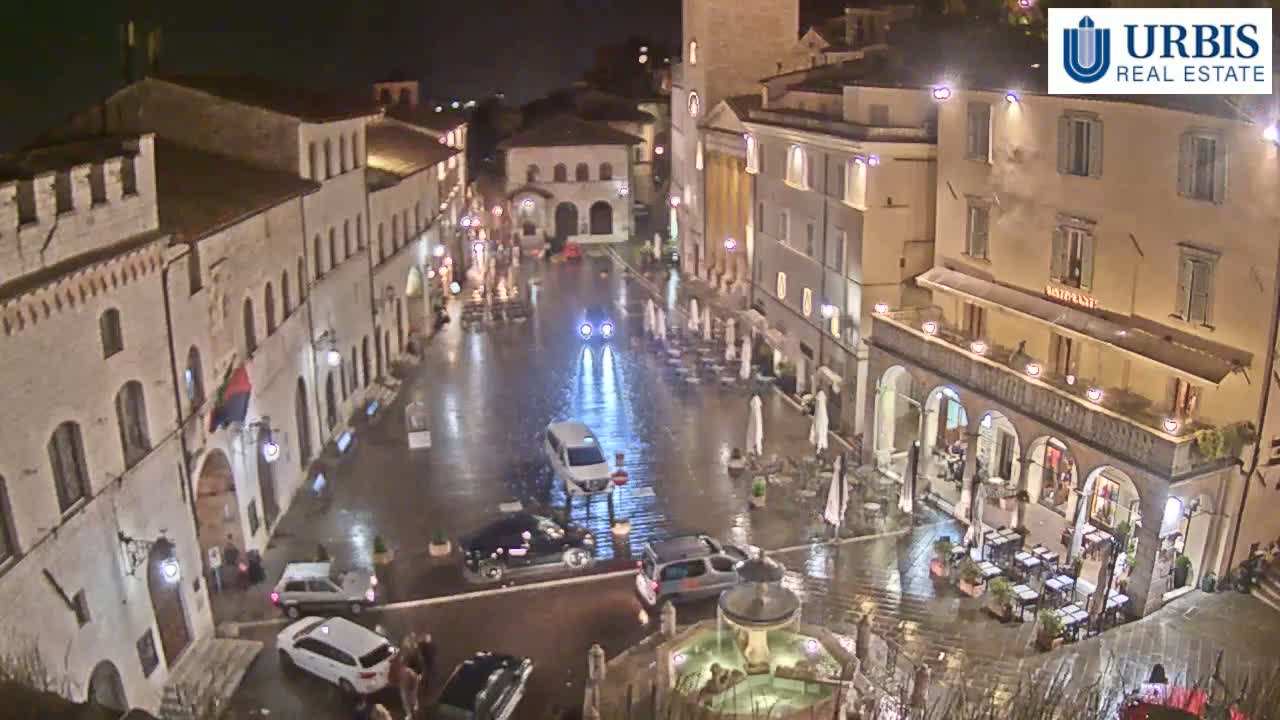 A rain-slicked town square at night is surrounded by illuminated buildings, with several cars parked and a few people walking around.