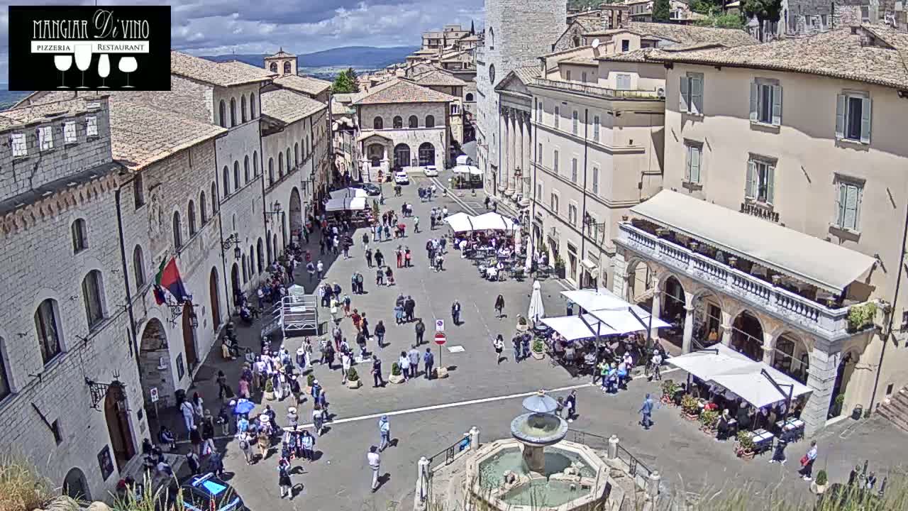 A sunny day in a town square, filled with people and featuring a fountain in the center, surrounded by buildings with outdoor seating.